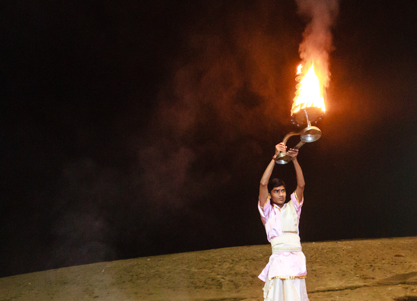 Against a background of the Ganges shore and a night sky, a young man in ceremonial attire holds a tower of flame aloft.